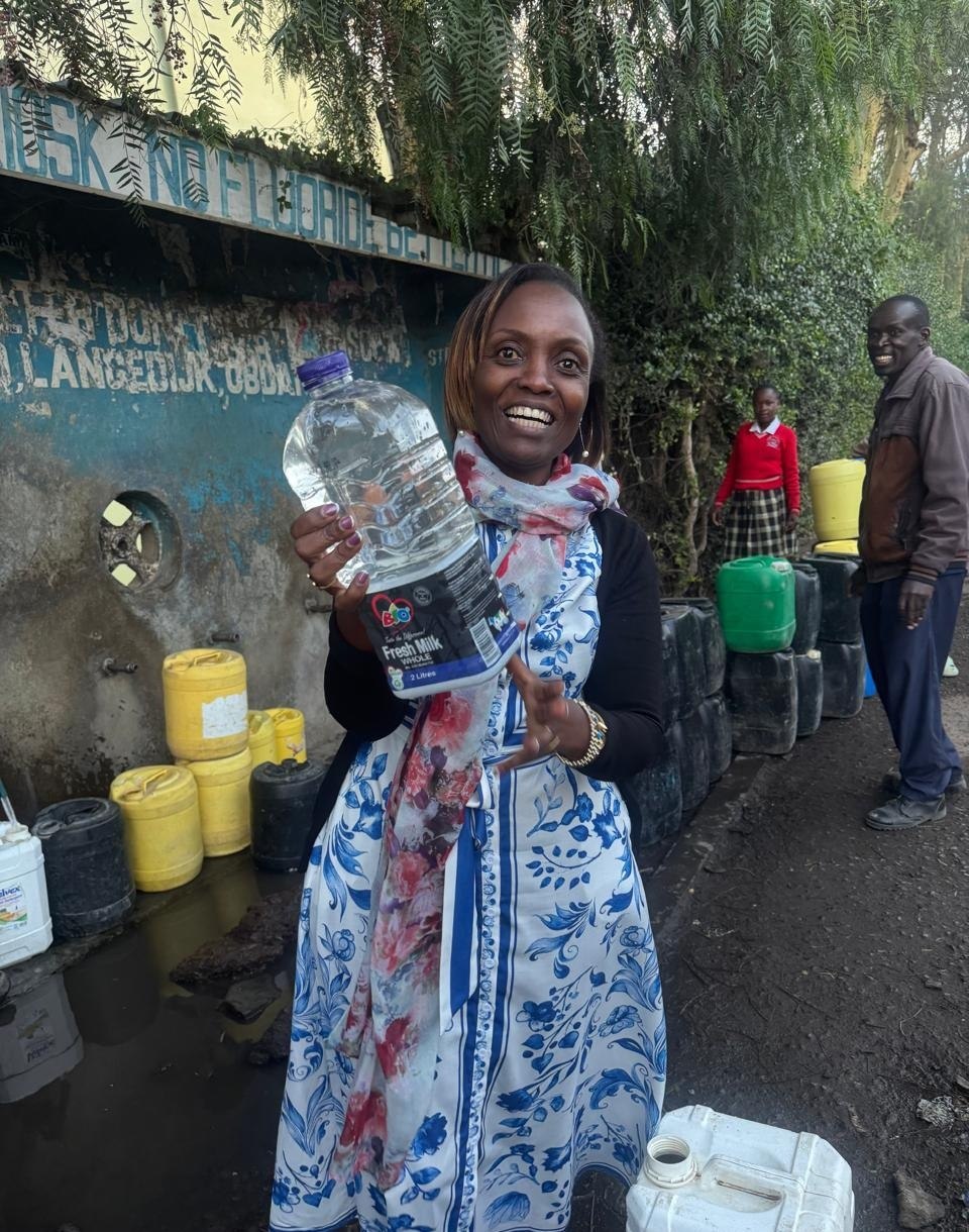 A woman holds up a bottle of clear water at a Kasarani water kiosk, jerry cans lined along the painted wall behind her