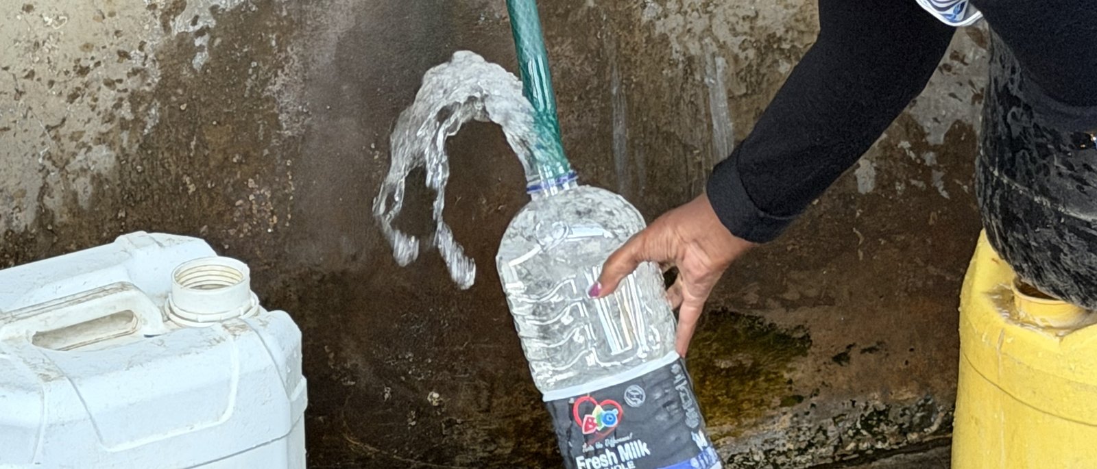 Clear water arcs from a hose into a repurposed bottle at a Kasarani water point, jerry cans waiting alongside