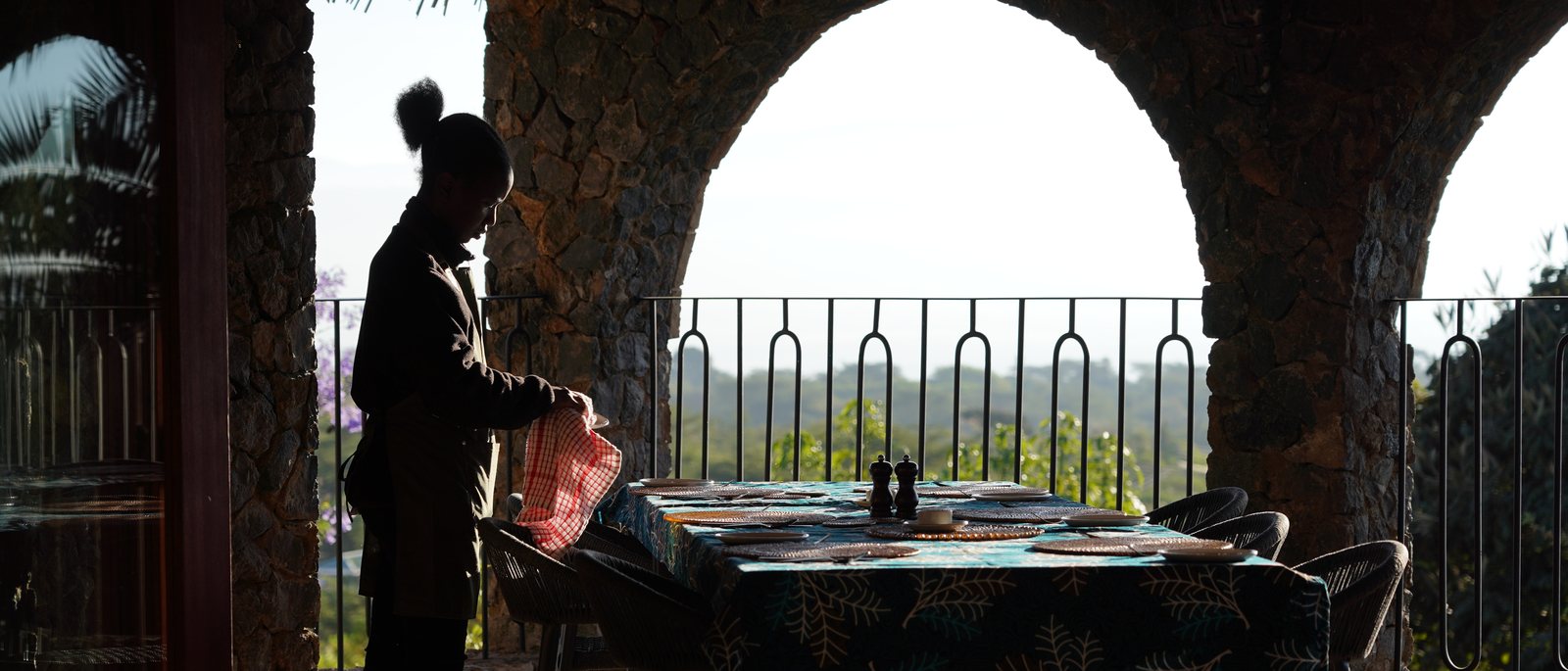 A staff member silhouetted against a stone arch, setting a long table above the Rift Valley landscape