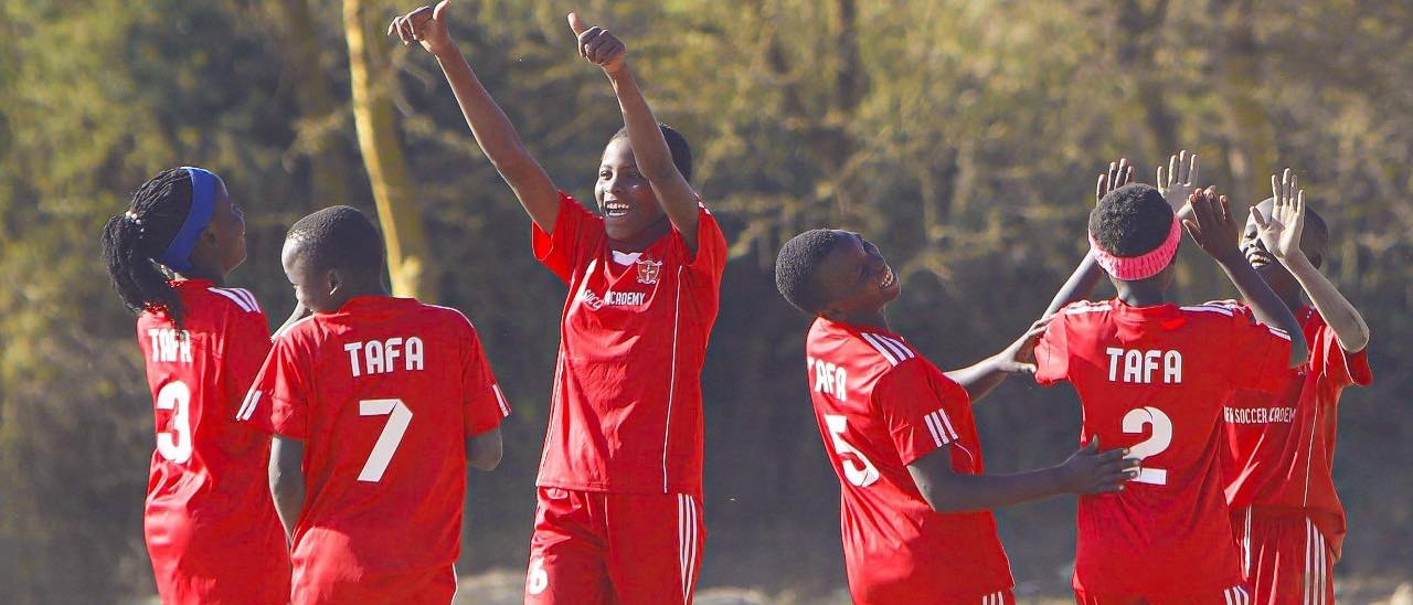 TAFA under-15 players in red kit celebrate together on the dry-grass pitch, dust in the late light