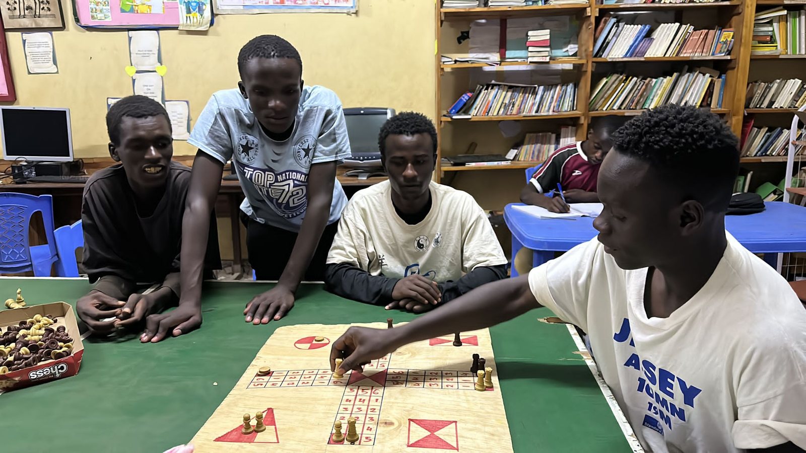 Young men around a board game in the TAFA community center library, bookshelves and a computer behind them