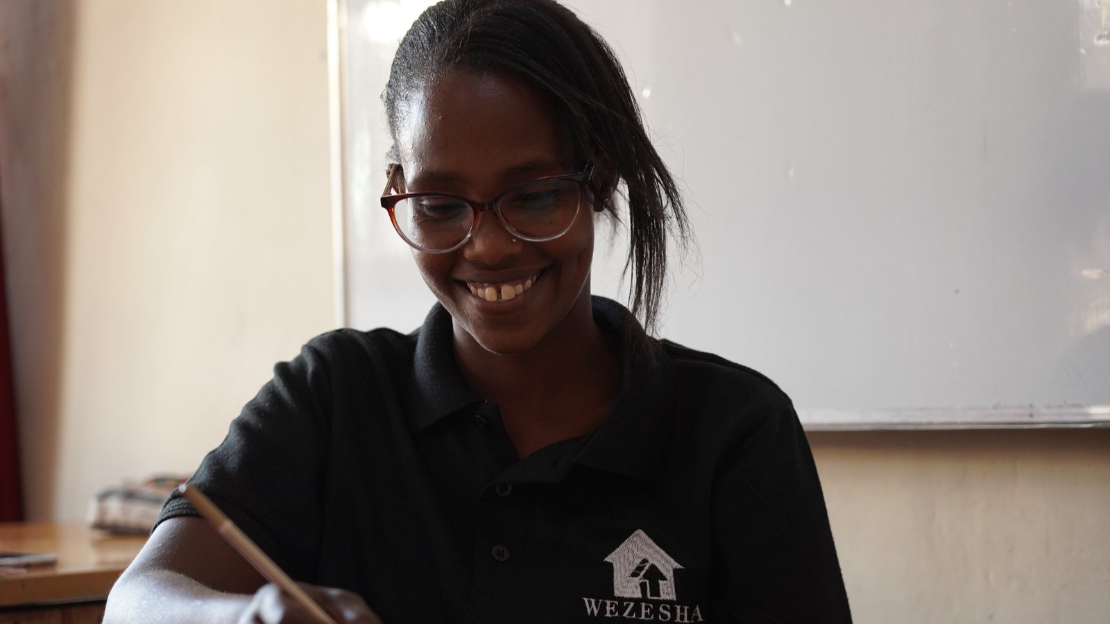 A young woman in a Wezesha polo smiles down at the notebook she is writing in